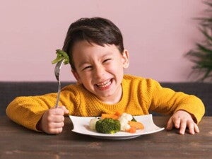 Menino sorridente segurando um garfo com brócolis e um prato cheio de vegetais cozidos à frente, representando alimentos para aumentar a imunidade de forma divertida e nutritiva.