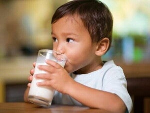 Criança pequena sentada à mesa e bebendo leite de um copo de vidro transparente. A foto tem foco no menino e no copo.