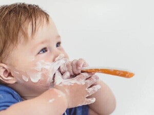 Close-up de um bebê, com o rosto e as mãos sujos de iogurte ou creme branco, segurando uma colher laranja e branca e olhando para cima.