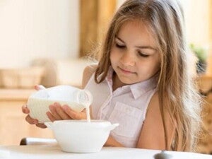 Menina sentada na mesa de jantar despejando leite em um tigela de cereais