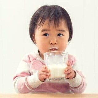 Bebê sentado à mesa, bebendo leite de um copo de vidro com bigode de leite no lábio superior. O fundo é branco.