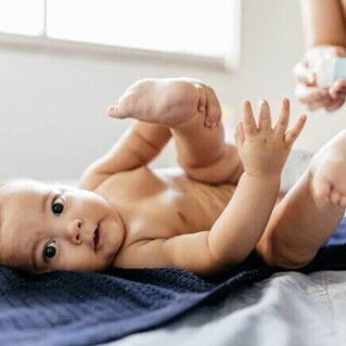 Um bebê deitado de costas em uma toalha azul escura, olhando para a câmera com as pernas levantadas e as mãos abertas, em um momento de troca de fraldas.