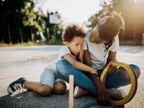Mãe e filho, uma criança de 6 anos, estão sentados no chão de um espaço ao ar livre. A mãe olha para o filho, que o abraça.