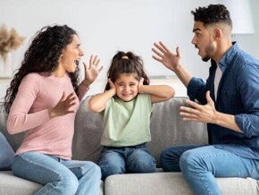 Pais discutindo sentados no sofá enquanto a criança, entre eles, cobre os ouvidos, demonstrando desconforto diante do conflito familiar.