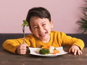 Menino sorridente segurando um garfo com brócolis e um prato cheio de vegetais cozidos à frente, representando alimentos para aumentar a imunidade de forma divertida e nutritiva.