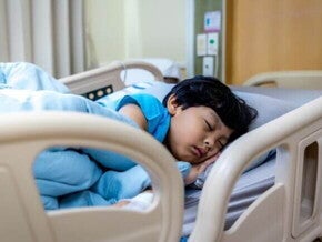 Uma criança dormindo profundamente em uma cama de hospital, enrolada em lençóis azuis, em um ambiente hospitalar.