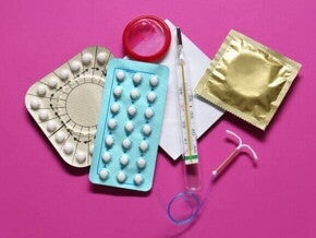 Visão aérea de vários métodos contraceptivos, incluindo cartelas de pílulas anticoncepcionais, preservativos (camisinhas), um DIU (dispositivo intrauterino) e um termômetro, dispostos sobre um fundo rosa.