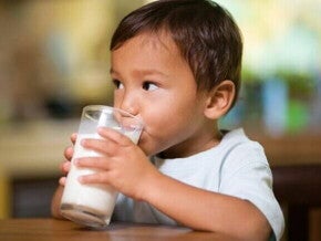Criança pequena sentada à mesa e bebendo leite de um copo de vidro transparente. A foto tem foco no menino e no copo.