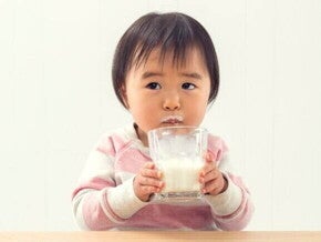 Bebê sentado à mesa, bebendo leite de um copo de vidro com bigode de leite no lábio superior. O fundo é branco.