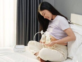 Uma mãe sorridente e sentada na cama utilizando uma bomba elétrica para extrair e coletar o leite materno em frascos.