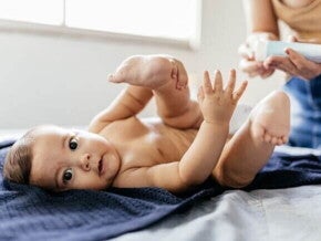 Assadura alergia alimentar Um bebê deitado de fralda em um trocador sobre uma toalha azul-escura, com as pernas levantadas. Na parte superior direita, mãos desfocadas de um adulto seguram um tubo de creme, preparando-se para aplicar pomada para assadura por alergia alimentar ou de contato.