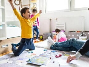 Sala com um adulto, onde só é possível ver suas pernas usando calça jeans e meia. Na mesma sala, estão três crianças. Uma garota com cerca de 1 ano de idade em segundo plano, branca, de cabelos curtos, de costas brincando com uma boneca. Outra garota com cerca de quatro anos, branca, cabelos loiros presos, calça jeans e blusa de manga comprida. Ela parece estar feliz dançando. A criança em destaque é um menino, branco, com cabelos curtos, de braços abertos comemorando uma vitória em um jogo 