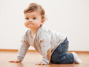 Um bebê engatinhando em um chão de madeira. Ele usa uma blusa cinza com cotoveleiras de camurça marrom e calça jeans.