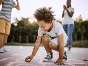 Imagem de uma criança ao centro brincando de uma brincadeira africana no chão. Ao fundo seus pais torcem por ela