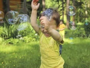 Um menino sorri com as mãos para o alto enquanto passa por bolhas de sabão em um ambiente externo arborizado, com outro menino ao fundo. A foto faz alusão ao tema de brincadeiras ao ar livre. 