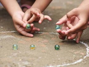 Duas crianças brincam de bola de gude no chão em um círculo desenhado de giz. A foto faz alusão ao tema de brincadeiras indígenas