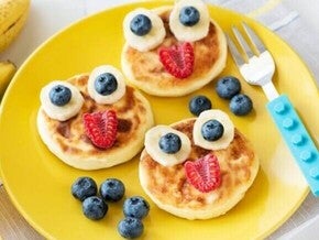 Três panquecas douradas decoradas de forma lúdica com fatias de banana, mirtilos e framboesas, formando rostos divertidos.