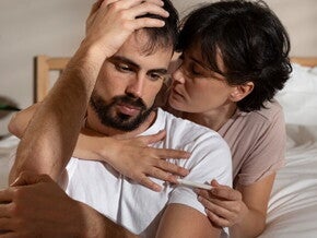 Casal sentado em uma cama, homem com semblante de preocupação recebendo abraço da mulher Casal sentado em uma cama, homem com semblante de preocupação recebendo abraço da mulher