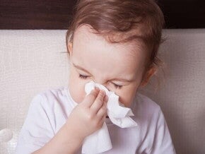 Bebê segura um lenço branco próximo ao nariz, sugerindo que está limpando secreção nasal, em um ambiente doméstico.