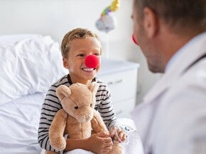 Um menino sorridente com um nariz de palhaço vermelho e segurando um ursinho de pelúcia está sentado em uma cama de hospital, olhando para um médico de costas para o observador. O médico também usa um nariz de palhaço vermelho. Um menino sorridente com um nariz de palhaço vermelho e segurando um ursinho de pelúcia está sentado em uma cama de hospital, olhando para um médico de costas para o observador. O médico também usa um nariz de palhaço vermelho.