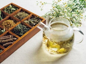 Com quantos meses o bebê pode tomar chá Uma chaleira de vidro com chá de ervas amarelado ao lado de uma caixa de madeira com compartimentos cheios de diversas ervas secas e flores brancas de camomila.