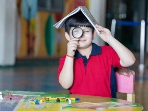 Um menino sorridente em uma mesa colorida, equilibrando um livro na cabeça e olhando através de uma lupa.