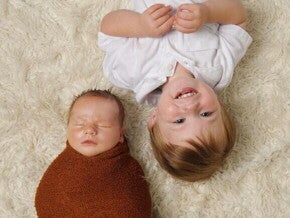 Uma foto de cima mostra um recém-nascido, envolto em um cobertor, dormindo ao lado de seu irmão mais velho, que está deitado de cabeça para baixo e sorrindo para a câmera. A imagem ilustra a relação inicial entre irmãos, abordando a temática da mudança de comportamento com a chegada do irmão mais novo.
