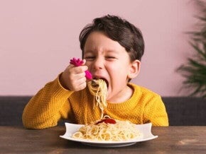 Criança sentada à mesa, comendo um prato de macarrão, em ambiente interno com parede rosa e planta ao fundo.