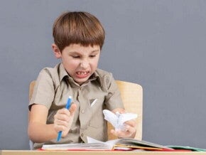 Menino em idade escolar sentado em uma carteira, com expressão de raiva ou frustração, cerrando os dentes e segurando um lápis ou caneta com força.