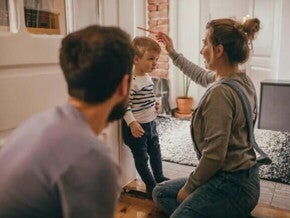 A mãe está ajoelhada checando o crescimento no filho, que está encostado na parede. Ela segura um lápis sobre a cabeça da criança para marcar a altura. O pai olha para a cena, um pouco mais distante. 