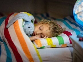 Criança dormindo tranquilamente abraçada a um bicho de pelúcia, coberta por um edredom listrado e colorido.