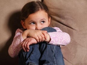 Uma criança medrosa sentada em um sofá marrom, abraçando os próprios joelhos com força e escondendo parte do rosto. Ela veste uma blusa rosa de manga longa e calça jeans, olhando para o lado com uma expressão de insegurança e apreensão.