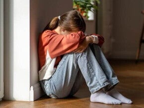 Uma jovem sentada no chão, com os braços abraçando os joelhos e a cabeça apoiada neles, demonstrando tristeza e isolamento.