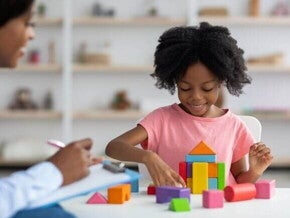 Uma menina sorrindo e organizando blocos coloridos, ao lado de uma mulher sentada com uma ficha na mão.