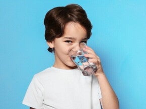 Um menino está bebendo água de um copo transparente, olhando para a câmera, sob um fundo azul.