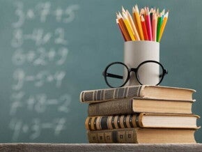 Uma pilha de livros sob uma mesa sustenta um par de óculos e uma caneca com lápis coloridos, com um quadro negro ao fundo