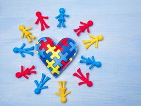 Coração feito de tecido com estampa de quebra-cabeça azul, vermelho e amarelo, simbolizando o autismo, cercado por pequenos bonecos coloridos de plástico.