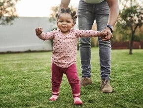 Imagem de um bebê cuja criação segue a abordagem de Emmi Pikler e que está brincando em um quintal supervisionado pela mãe que está lhe dando a mão.