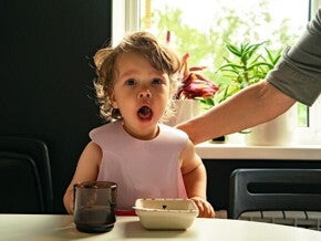 Um bebê com babador sentado à mesa, com a boca aberta, olhando diretamente para a câmera, com a mão de um adulto ao fundo. Um bebê com babador sentado à mesa, com a boca aberta, olhando diretamente para a câmera, com a mão de um adulto ao fundo.