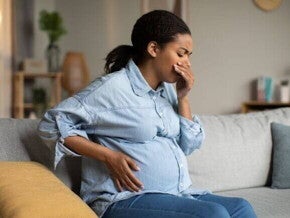 Gestante sentada no sofá com a mão na barriga e cobrindo a boca, demonstrando mal-estar, cenário comum para quem procura saber o que é bom para enjoo na gravidez.