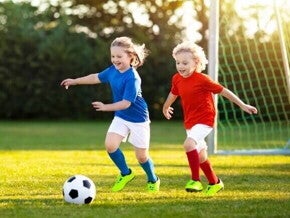 Duas crianças jogando futebol em um campo gramado sob a luz do sol, representando a prática de esportes para crianças.