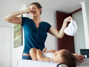 Uma mulher em pé, segurando uma fralda em uma mão e tapando o nariz com a outra mão, com um bebê deitado em sua frente