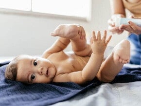 Um bebê deitado de costas em uma toalha azul escura, olhando para a câmera com as pernas levantadas e as mãos abertas, em um momento de troca de fraldas.