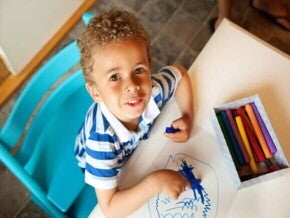 criança sentada em cadeira e mesa infantil com giz de cera azul em mão, desenhando em folha de papel, outras cores de giz disponíveis sobre a mesa criança sentada em cadeira e mesa infantil com giz de cera azul em mão, desenhando em folha de papel, outras cores de giz disponíveis sobre a mesa