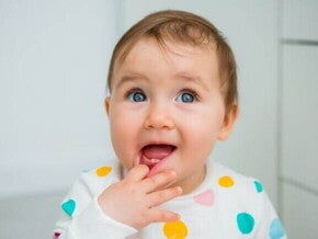 Bebê sentado, levando a mão à boca, em um ambiente interno bem iluminado.