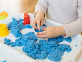 Close das mãos de uma criança interagindo com areia cinética azul em uma bandeja branca.