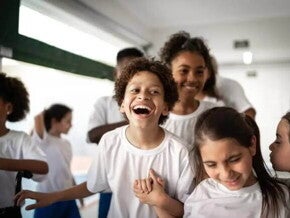 Imagem de um ambiente escolar com várias crianças vestidas de uniforme. O destaque está para um menino gargalhando, bem no centro da foto, de mãos dadas com uma menina