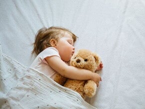 Uma bebê deitada em uma cama, dormindo, abraçada com um ursinho de pelúcia.