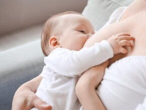 Bebê de roupa branca sendo amamentado pela mãe sentada em um sofá