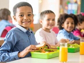 Crianças felizes e sorridentes, comendo sanduíches e suco durante o recreio, utilizando uma lancheira verde cheia de alimentos saudáveis como vegetais e frutas.